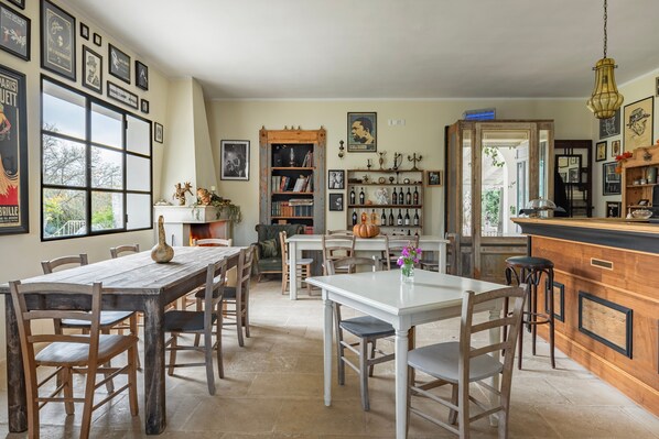 A dining area with a large window, a wooden table, and chairs. There is a bookshelf with books and decorative items, and a kitchen area with a bar counter. - Room 'Dalia 2' with Shared Terrace, Wi-Fi and Air Conditioning (Ortelle)