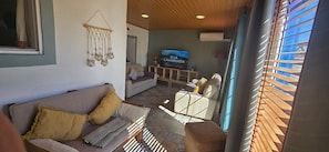 A living room with a sofa, cushions, a TV, a wooden shelf, and a window with blinds.