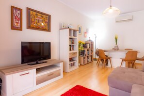 Een moderne woonkamer met een flatscreen televisie, een witte tv-meubel, een boekenkast, een eethoek met stoelen, en een rode vloerkleed.