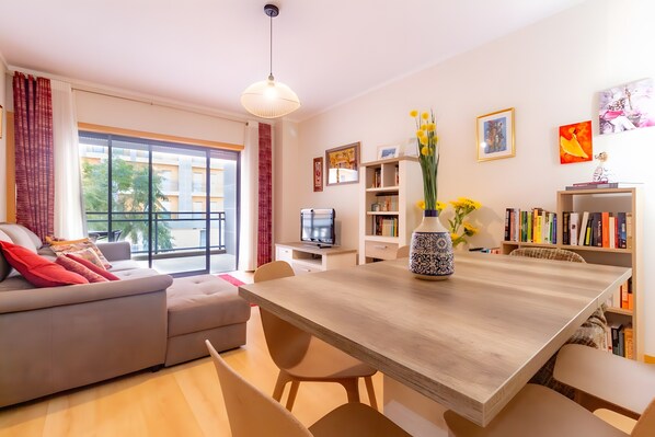 A modern living room with a sofa, dining table, chairs, a TV, bookshelves, and artwork. - Apartment 'Marina Formosa' with Shared Pool, Wi-Fi and Air Conditioning (Olhão)