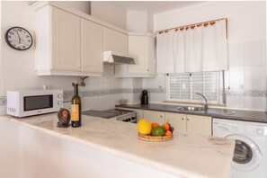 Una cucina con mobili bianchi, un forno a microonde, un lavello, una lavatrice e una ciotola di frutta sul piano di lavoro.