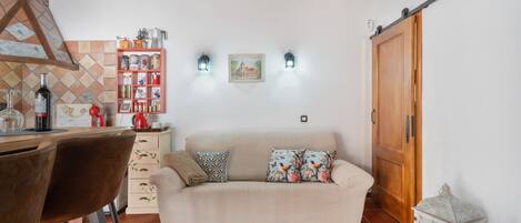 A cozy living area with a sofa, a wooden ceiling, a kitchen area with a shelf of items, and a sliding door.