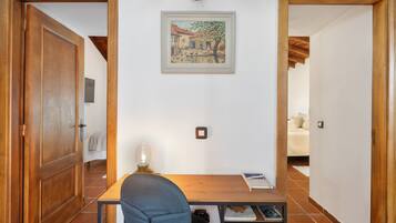 Una stanza con una scrivania in legno, una sedia, una lampada, un quadro appeso al muro e un letto sullo sfondo.