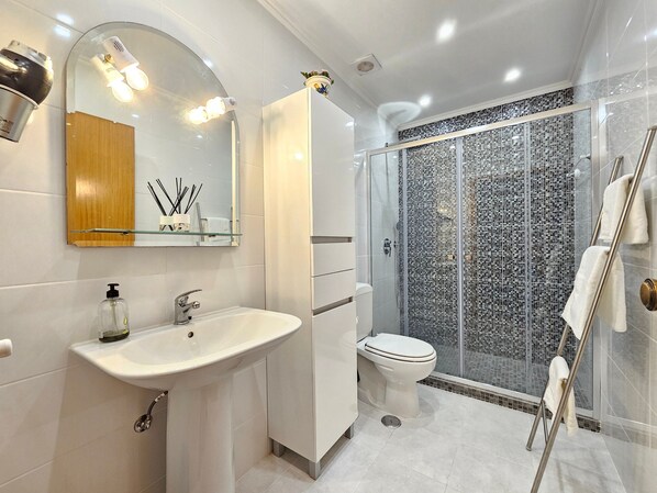 A modern bathroom with a white sink, a toilet, a glass shower enclosure, and a mirror with lights.