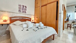 Ein ordentlich bezogenes Bett mit weißer Decke, ein Holzschrank und ein kleiner Tisch mit Lampe.