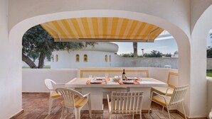 An outdoor dining area with a table set for four, surrounded by white chairs, under a striped awning. - Apartment 'Sail Golf Vilamoura' with Shared Pool, Wi-Fi and Air Conditioning (Quarteira)