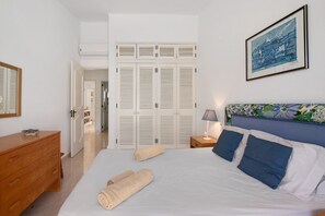 Une chambre à coucher comprenant un lit, une commode, une table de chevet avec une lampe, un tableau au mur et un ensemble de portes à persiennes blanches.