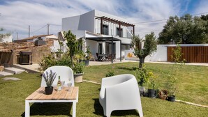 Una casa moderna con un patio, sillas blancas, una mesa con una botella y un vaso, y plantas en macetas.