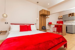 Una moderna camera d'albergo con un letto grande, una testiera in legno, una parete di colore rosso e un angolo cottura, oltre a una lampada a sospensione.