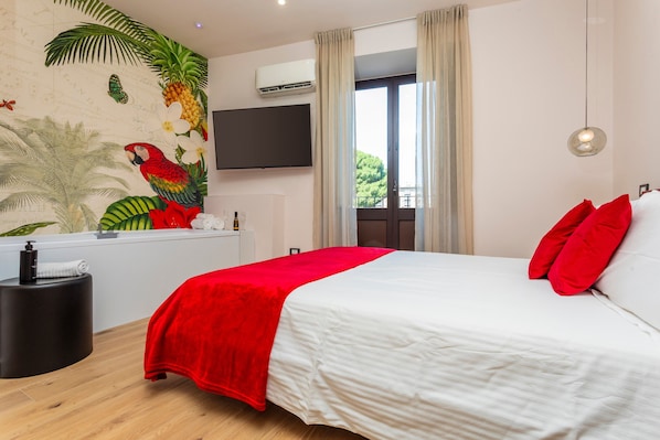 Una camera da letto con un letto, cuscini rossi, una TV a muro, un condizionatore d'aria fissato al muro e una finestra con tende.