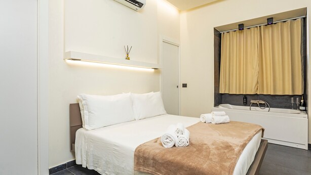 A hotel room with a bed, a bathroom with a bathtub, a window with curtains, and a wall-mounted air conditioner.