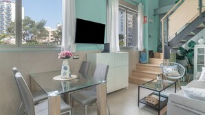 A modern living room with a glass dining table, a sofa, a TV mounted on the wall, and a staircase leading to an upper floor.