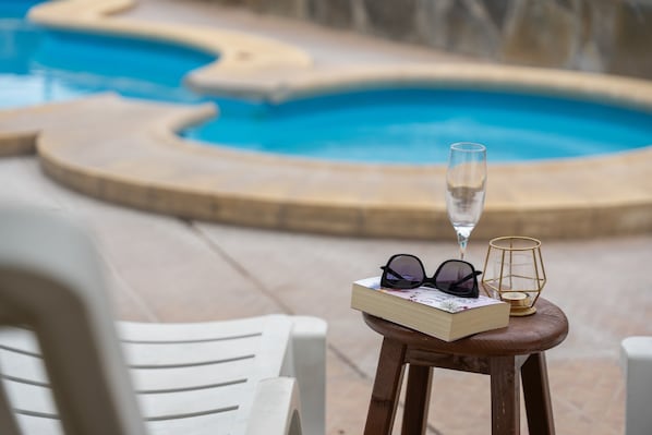 Escena junto a la piscina con un libro, gafas de sol y una copa de champán sobre un taburete de madera.