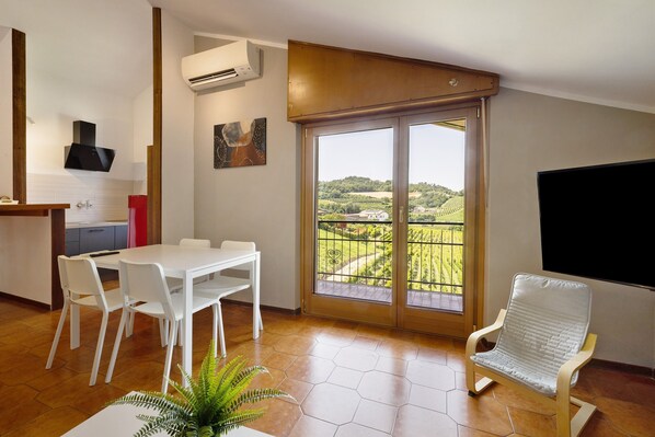 Una cocina moderna con armarios blancos, un comedor con mesa y sillas blancas, un balcón con vistas a zonas verdes, y un televisor de pared.