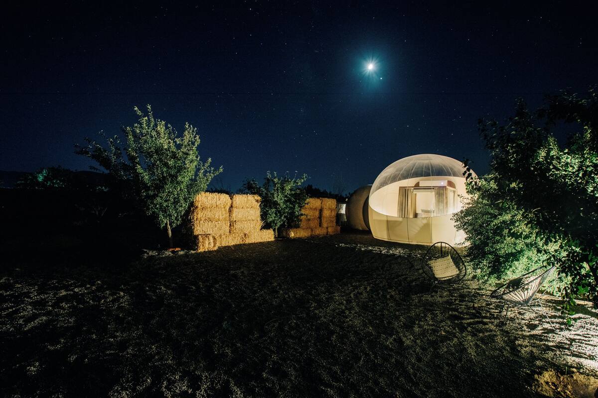 A dome-shaped building with a door, surrounded by trees and hay bales, under a starry night sky.