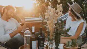 Two people sitting at an outdoor table with a tray of bottles and glasses, surrounded by plants and a bench.
