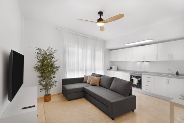 A modern living room with a sectional sofa, a flat-screen TV, a ceiling fan, a potted plant, and a kitchen area with white cabinets and appliances.