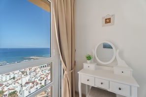 Una habitación con tocador blanco, espejo, una planta pequeña y vistas al mar y edificios.