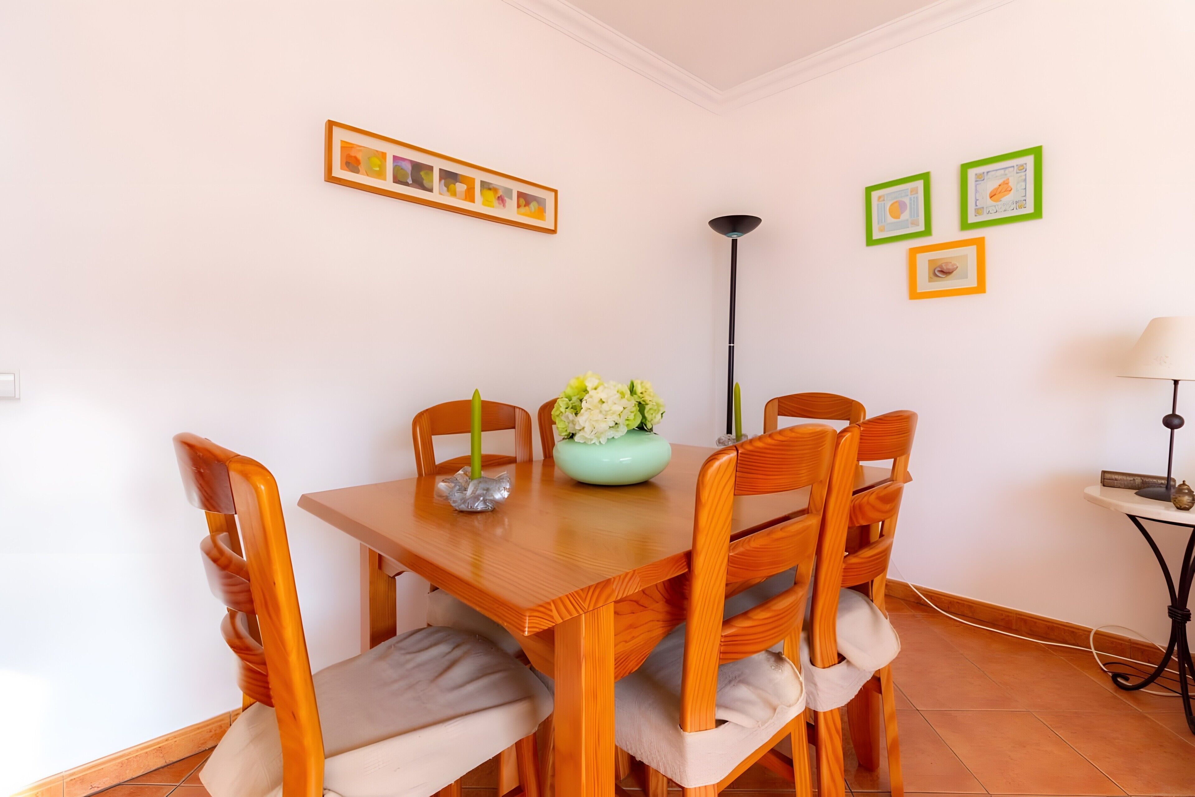 Uma área de refeições com mesa e cadeiras de madeira, um vaso com flores e quadros nas paredes.