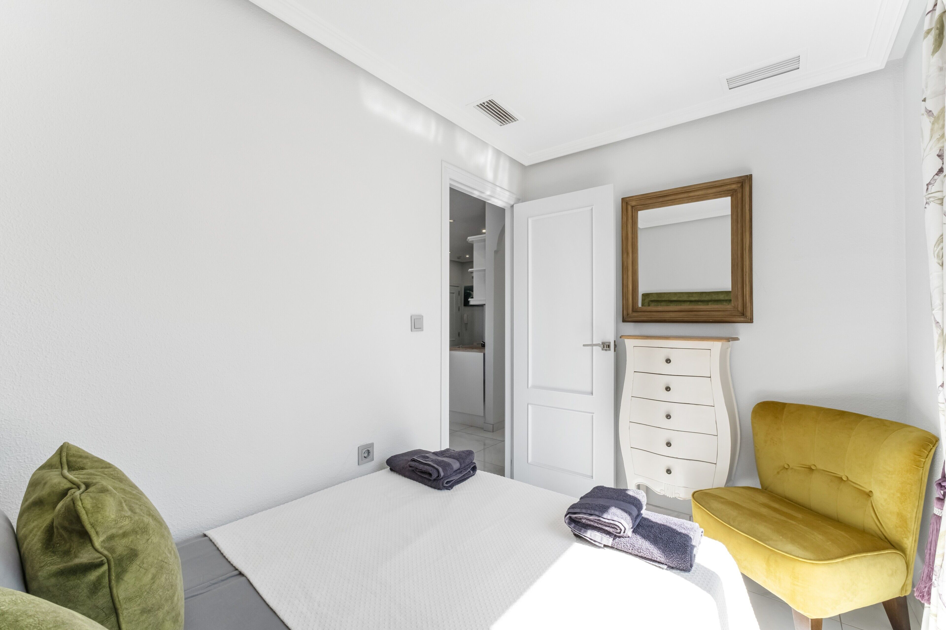 A bedroom with a bed, a dresser, a chair, a mirror, and a door leading to a bathroom.