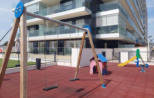 Ein Spielplatz mit Schaukeln und einer Rutsche, umgeben von einem roten Kachelboden und einem modernen Gebäude mit Balkonen.