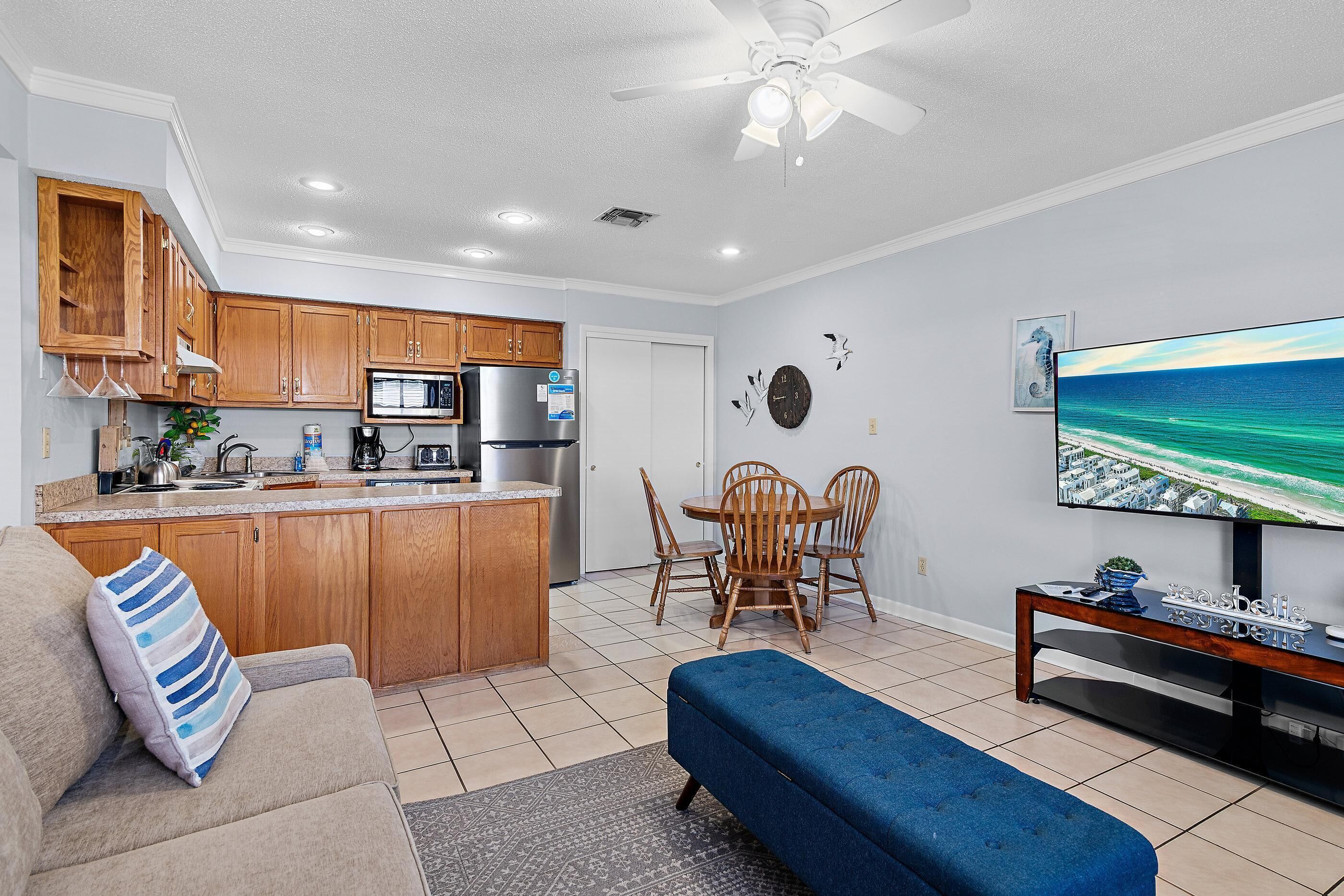 A modern kitchen with wooden cabinets, a central island, and a dining area with a TV displaying a beach scene.