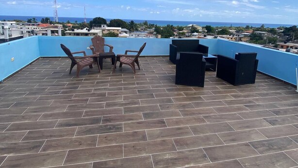 A rooftop terrace with a blue railing, a table with chairs, and a view of the ocean and cityscape.