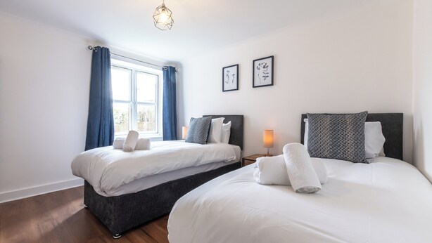 Hotellihuone, jossa on kaksi sänkyä, sinisillä verhoilla varustettu ikkuna, yöpöytälamppu ja seinällä kehystetty taideteos.