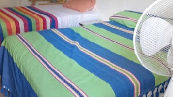 Ein Zimmer mit zwei Betten, jeweils mit bunter, gestreifter Bettwäsche und einem Kopfkissen. Ein weißer Ventilator steht auf dem Boden neben einem Bett.