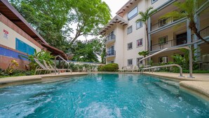 A swimming pool with a metal ladder, surrounded by greenery and a multi-story building with balconies.