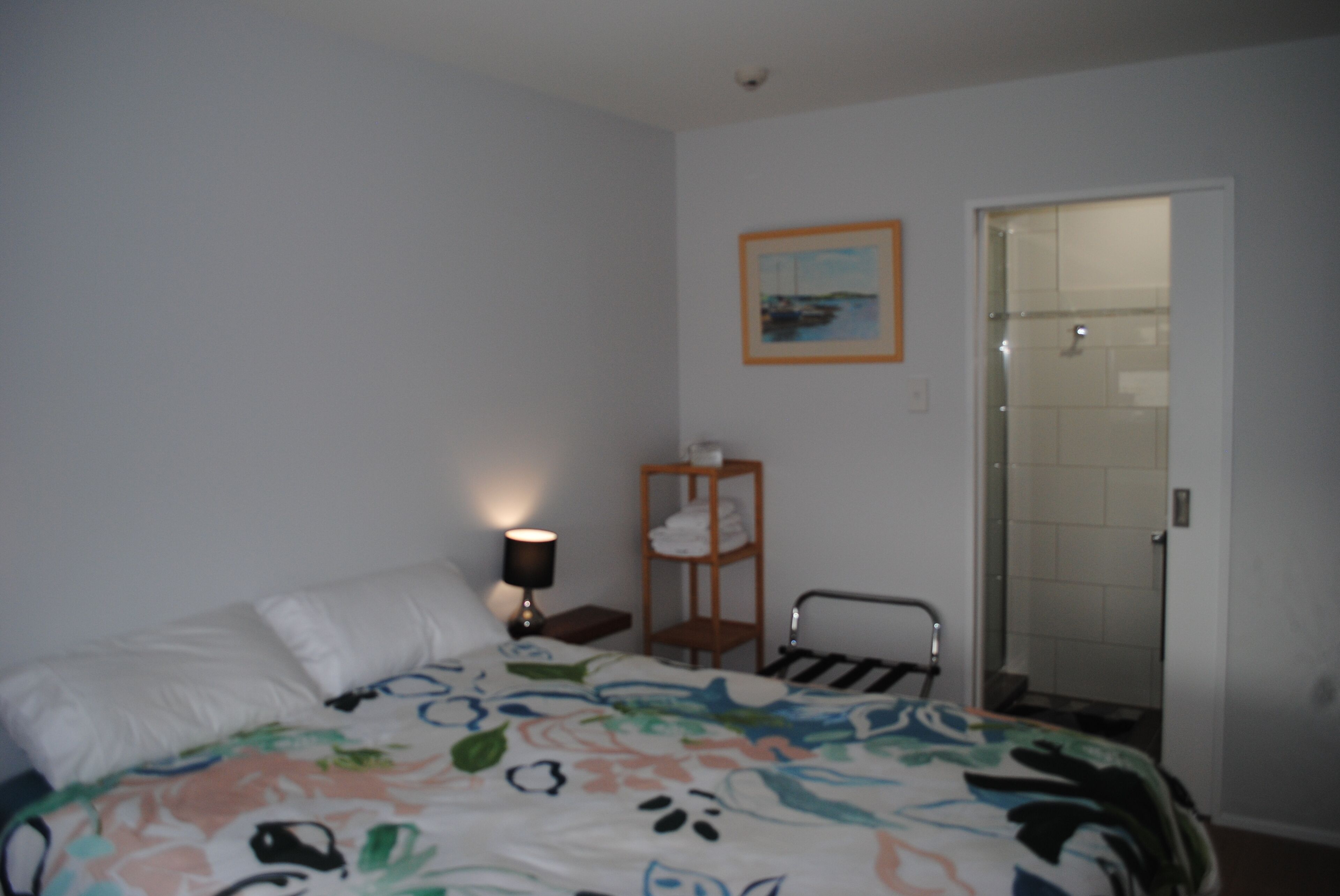 A bedroom with a bed, a nightstand with a lamp, a shelf with towels, and a door leading to a bathroom.