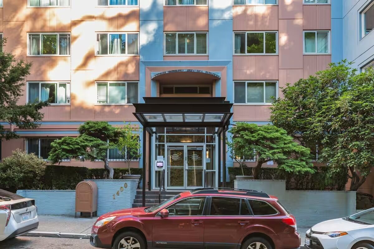Et hotel med flere etager, en central indgang, et overdækket venteområde og en parkeringsplads med biler.