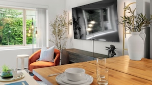 Un soggiorno moderno con un tavolo da pranzo in legno apparecchiato per due persone, un divano, una TV a parete e una grande finestra con vista su piante.