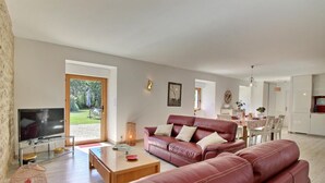 A modern living room with a leather sofa, a coffee table, and a TV. There is a dining area with a table and chairs, and a kitchen in the background.