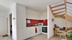 A compact kitchen with white cabinets, a red accent wall, a microwave, a kettle, and a wooden chair.