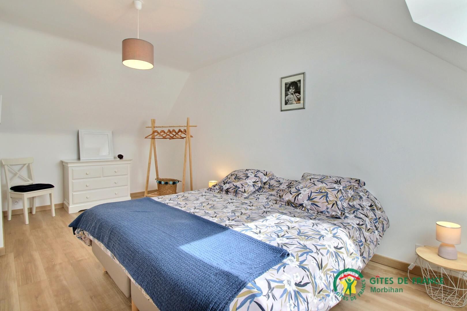 A bedroom with a bed, a desk, a chair, a hanging laundry rack, and a small bedside table with a lamp.