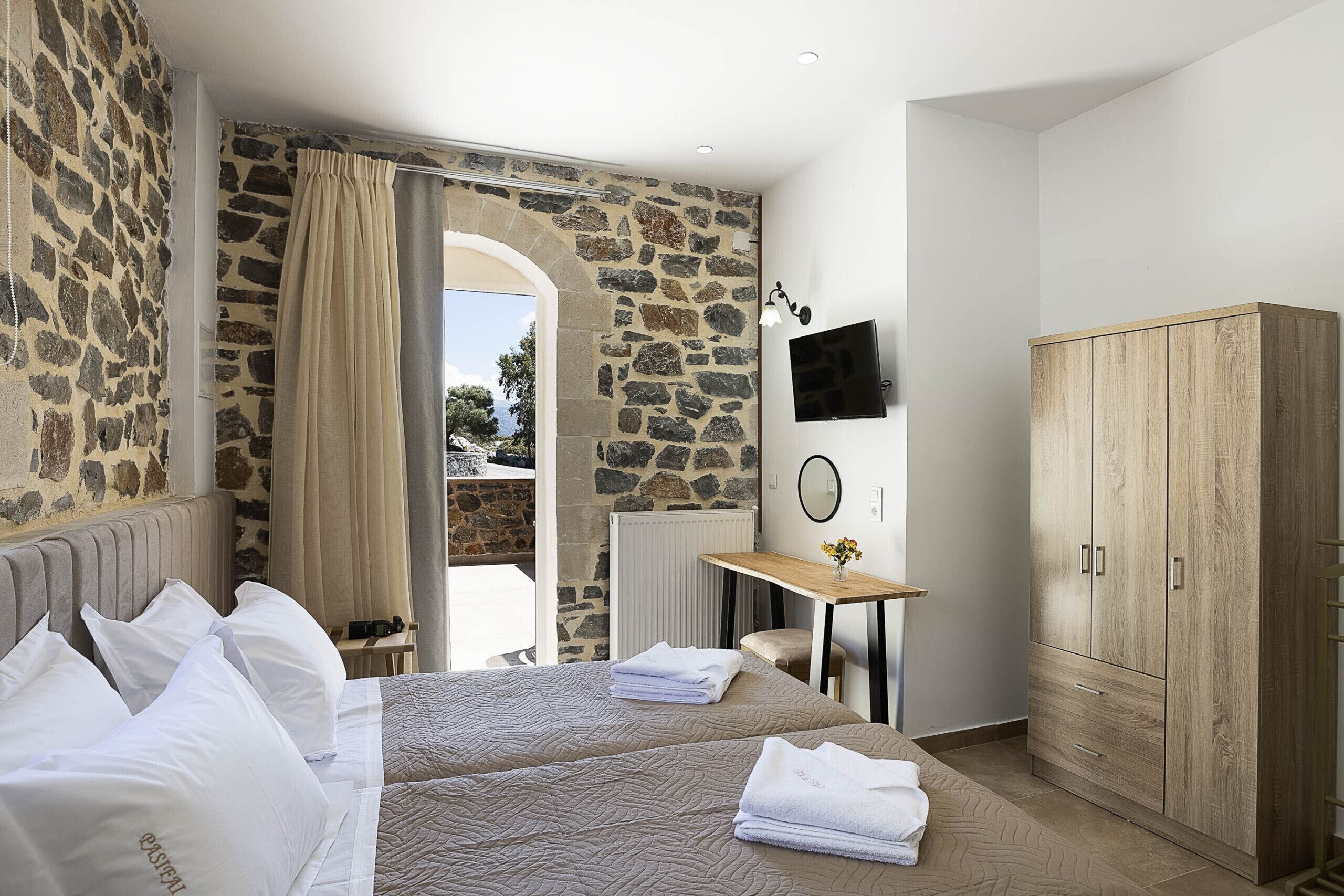 Une chambre d’hôtel avec deux lits, un mur en pierre, une armoire en bois, un bureau avec une chaise et un téléviseur.