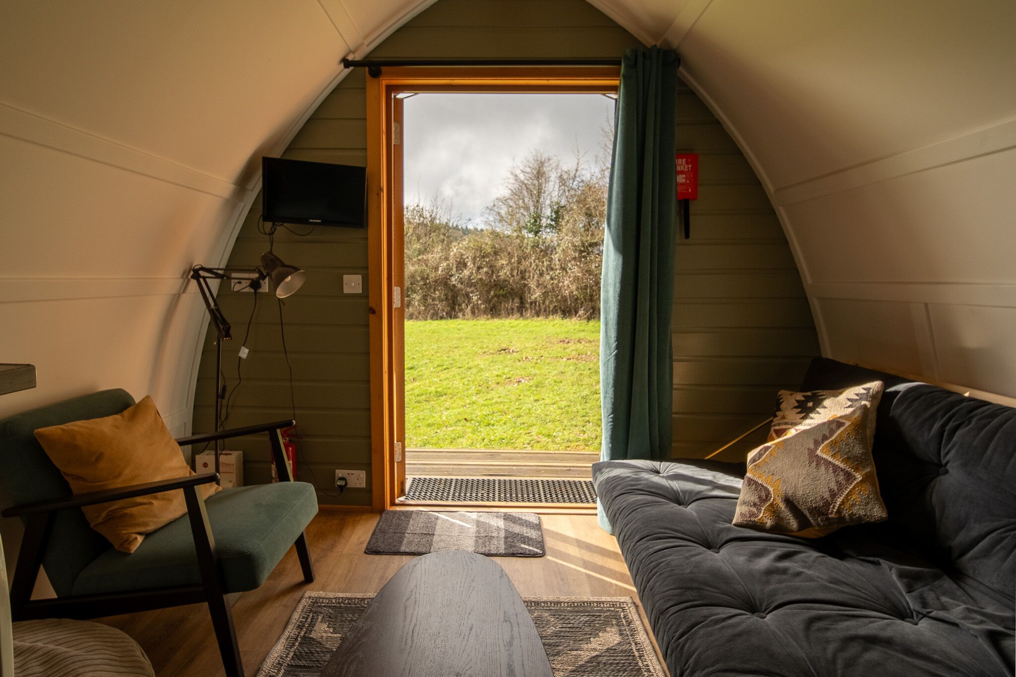 Une pièce confortable avec un canapé, un fauteuil, un téléviseur fixé au mur et une grande fenêtre donnant sur un espace verdoyant.