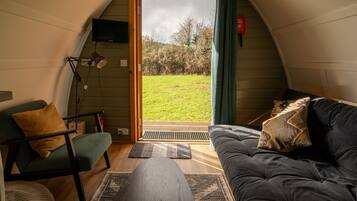 Une pièce confortable avec un canapé, un fauteuil, un téléviseur fixé au mur et une grande fenêtre donnant sur un espace verdoyant.