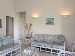Ein Zimmer mit einem weißen Sofa, einem weißen Sessel, einem Glastisch und einem kleinen Tisch mit einer Lampe. Eine Tür führt in einen anderen Raum, und ein Gemälde hängt an der Wand.