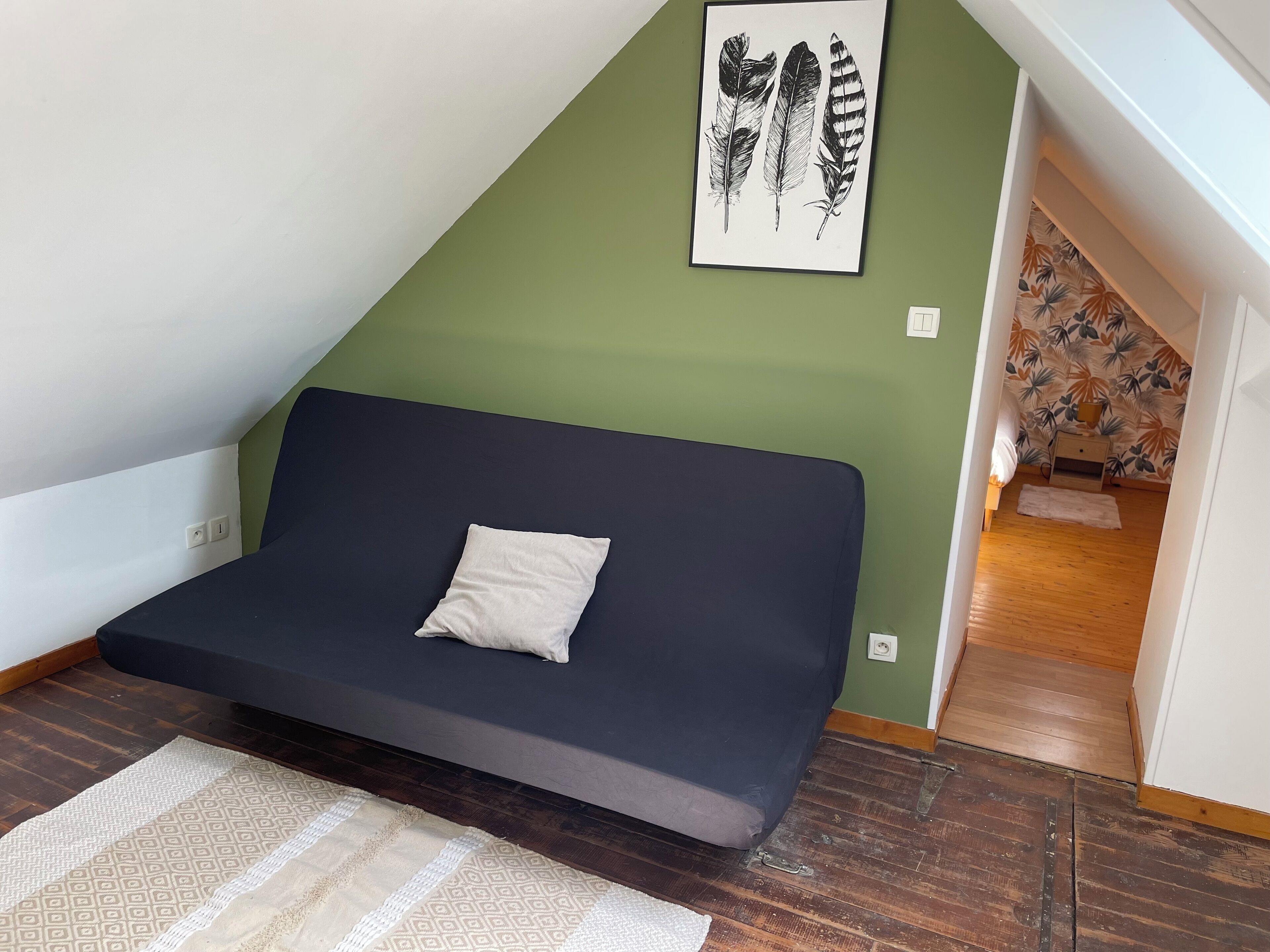 Une petite pièce accueillante, avec un canapé bleu foncé, un coussin blanc et une œuvre d’art représentant des plumes, encadrée, accrochée au mur vert.