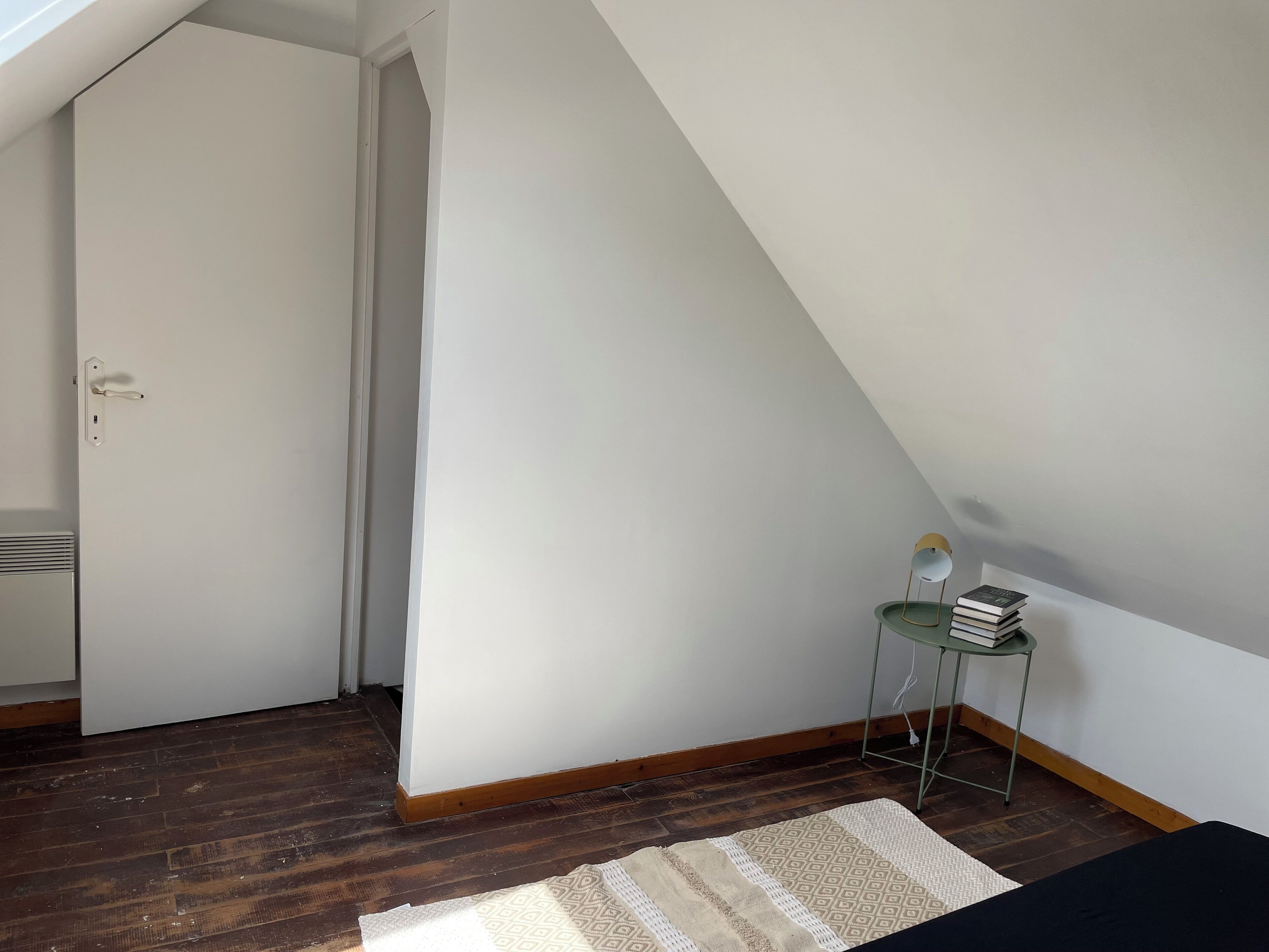 A room with a sloped ceiling, a door, a small table with books, a patterned rug, and wooden flooring.