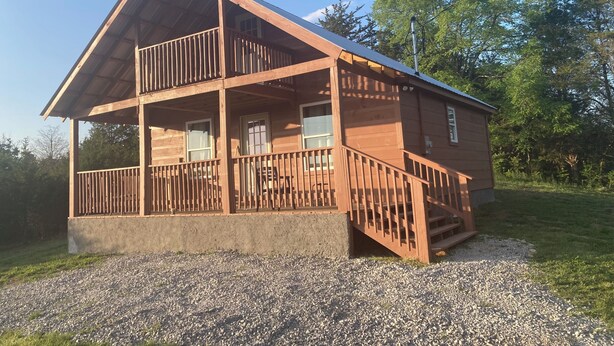 A two-story wooden cabin with a balcony on the upper floor, a gravel driveway, and surrounded by trees.