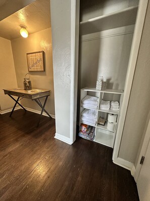 Une pièce avec un plancher en bois, un bureau avec une lampe, une étagère avec des serviettes et une armoire avec des vêtements suspendus.
