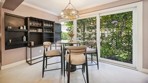 A dining area with a round table, four chairs, a chandelier, and a large window with a view of greenery.