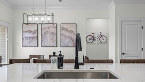 Una cocina moderna con encimera de mármol blanco, pileta de acero inoxidable y grifo negro. Se observan tres obras de arte enmarcadas en la pared, una bicicleta y una puerta al fondo.