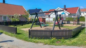 A playground with swings and a slide, surrounded by a wooden fence and houses with red roofs.