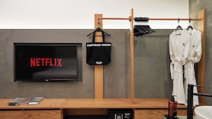 Un baño moderno con un tocador de madera, un bolso negro colgado en un perchero, una bata blanca en un perchero y un televisor mostrando el logotipo de Netflix.