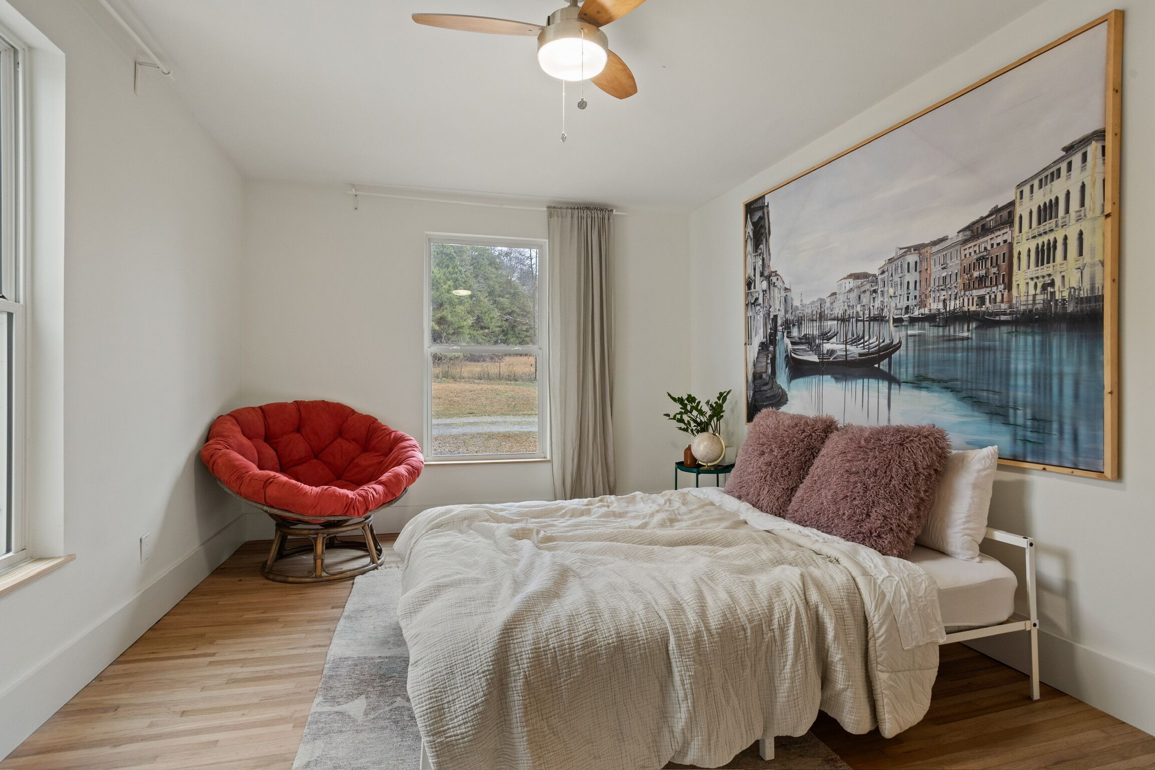 Un dormitorio con una cama, una silla roja, un ventilador de techo, una ventana y un cuadro grande que muestra un canal.
