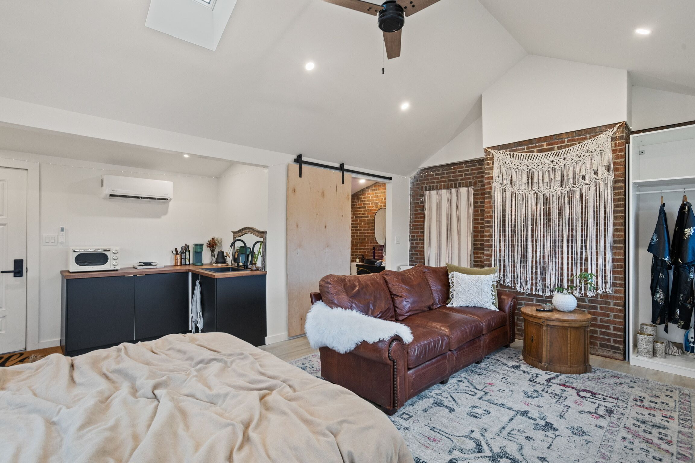 Quarto moderno com cama, um sofá de couro marrom, mesa de cabeceira de madeira pequena e um pendurar de macramé na parede.